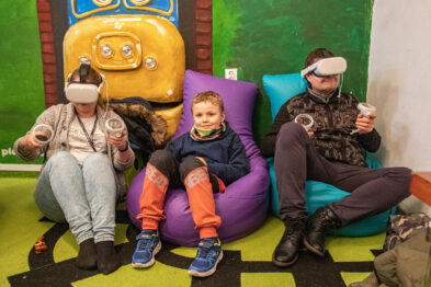 Jest to zdjęcie trzech osób siedzących na kolorowych pufach i korzystających z gogli wirtualnej rzeczywistości. Dwóch dorosłych i jedno dziecko, które trzymają w dłoniach kontrolery. W tle widoczne są elementy wystroju nawiązujące do tematyki kolejowej, w tym żółta dekoracja z kółkiem i sygnałami świetlnymi przypominającymi pociąg.