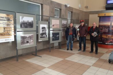 Na zdjęciu widoczna jest ekspozycja fotografii umieszczona na metalowych, mobilnych panelach w przestrzeni dworca kolejowego. Przedstawione prace to czarno-białe zdjęcia o tematyce kolejnictwa. Cztery osoby oglądają wystawę, a w tle widoczny jest fragment wnętrza dworca z jasną posadzką i informacyjnymi elementami.