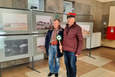 Na zdjęciu dwaj mężczyźni stoją w pomieszczeniu z wystawą fotografii. Za nimi widać plansze z czarno-białymi i kolorowymi zdjęciami kolejowymi umieszczone na metalowych stojakach. W tle dostrzec można elementy wnętrza dworca kolejowego, w tym kolumny i błyszczącą posadzkę.