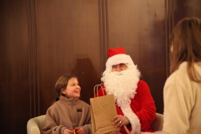 Na zdjęciu widać osobę przebraną za Świętego Mikołaja, trzymającą brązową papierową torbę, która jest wręczana uśmiechniętemu dziecku. Osoba w kostiumie Mikołaja i dziecko są wewnątrz pomieszczenia z brązowymi ścianami oraz dają wrażenie ciepłej, radosnej atmosfery. Po prawej stronie fotografii pojawia się część twarzy innej młodej osoby, która obserwuje interakcję.
