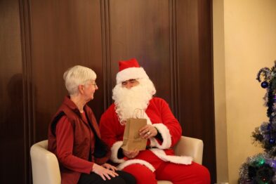Na fotografii widoczna jest starsza pani siedząca obok postaci w przebraniu Świętego Mikołaja. Obydwoje znajdują się w pomieszczeniu o ciepłej atmosferze, a Mikołaj trzyma w ręce prezent. W tle delikatnie zaznacza się dekoracja świąteczna, co podkreśla temat wydarzenia mikołajkowego.