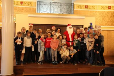 Na zdjęciu widać grupę uśmiechniętych dzieci ze szkoły podstawowej i seniorów zgromadzonych w eleganckim wnętrzu, pozujących do wspólnego zdjęcia. W centrum stoi postać ubrana w strój Świętego Mikołaja, która jest otoczona przez uczestników wydarzenia. Atmosfera wydaje się radosna, a pomieszczenie jest jasne i przytulne, co podkreśla miły charakter spotkania międzypokoleniowego.