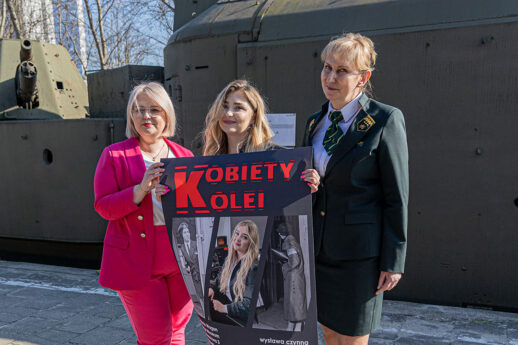 Na tle pociągu pancernego pozują trzy kobiety: kobieta w czerwonym ubraniu biurowym ,młoda kobieta i kobieta w mundurze Kolei Mazowieckich. Młoda kobieta trzyma plakat wystawy, na którym jest jej fotografia