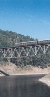 Most kolejowy na rzece. Na moście widoczny parowóz z dwoma wagonami pasażerskimi. Okolica górzysta