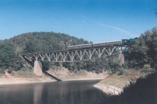 Most kolejowy na rzece. Na moście widoczny parowóz z dwoma wagonami pasażerskimi. Okolica górzysta