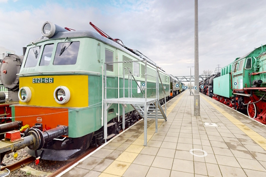 Muzealny peron. Po lewej zielona lokomotywa z żółtym czołem. Po prawej widać fragment zielonego parowozu z czerwonymi kołami