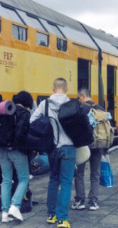 Podróżni z plecakami turystycznymi i torbami na peronie. Obok stoi żółty wagon piętrowy.