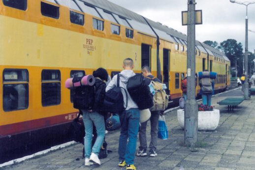 Podróżni z plecakami turystycznymi i torbami na peronie. Obok stoi żółty wagon piętrowy.