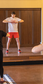 Na scenie młody mężczyzna w stroju sportowym pokazuje ćwiczenia. Na sali trzy starsze kobiety ćwiczą
