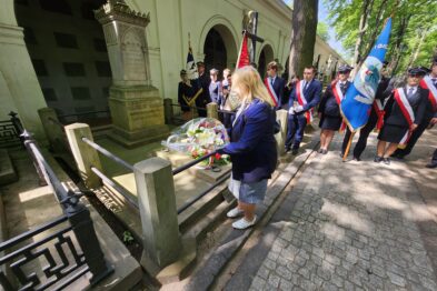 Kobieta składająca wiązankę kwiatów na grobie. Wokół poczty ze sztandarami, przepasani biało-czerwonymi wstęgami