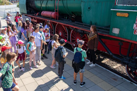 Grupa dzieci na peronie słucha przewodnika, który stoi przy zielonym parowozie
