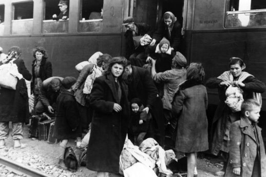 Zdjęcie czarno-białe. Na pierwszym planie młoda kobieta patrzy na fotografa robiącego to zdjęcie. Za nią tłum ludzi z walizkami i tobołkami wysiadający z pociągu