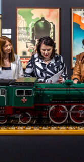 Trzy osoby siedzą przy stole w muzeum, podpisując dokumenty. Przed nimi stoi szczegółowy model zielonego parowozu. W tle widoczne są plakaty kolejowe oraz ekspozycja muzealna