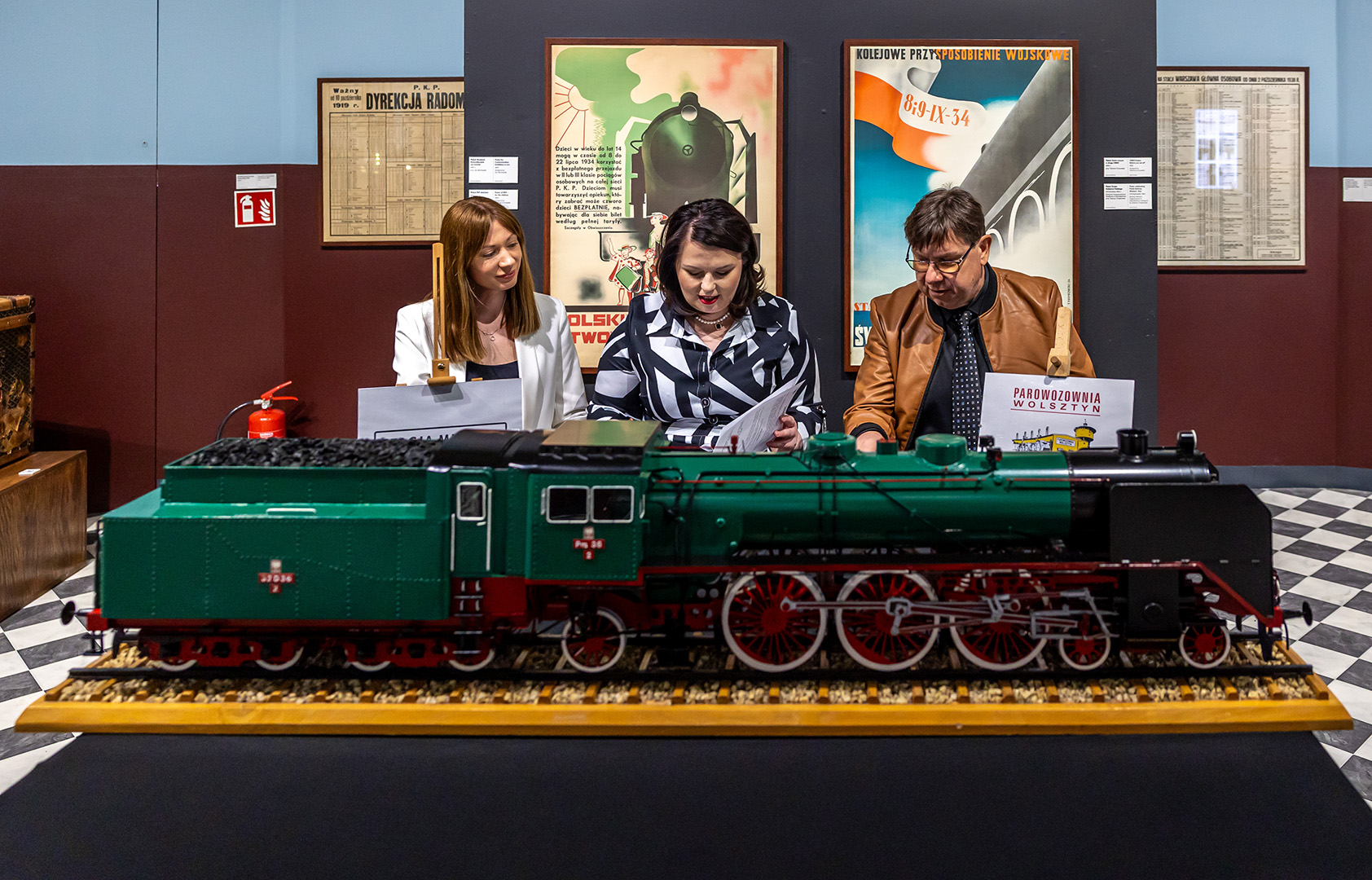 Trzy osoby siedzą przy stole w muzeum, podpisując dokumenty. Przed nimi stoi szczegółowy model zielonego parowozu. W tle widoczne są plakaty kolejowe oraz ekspozycja muzealna