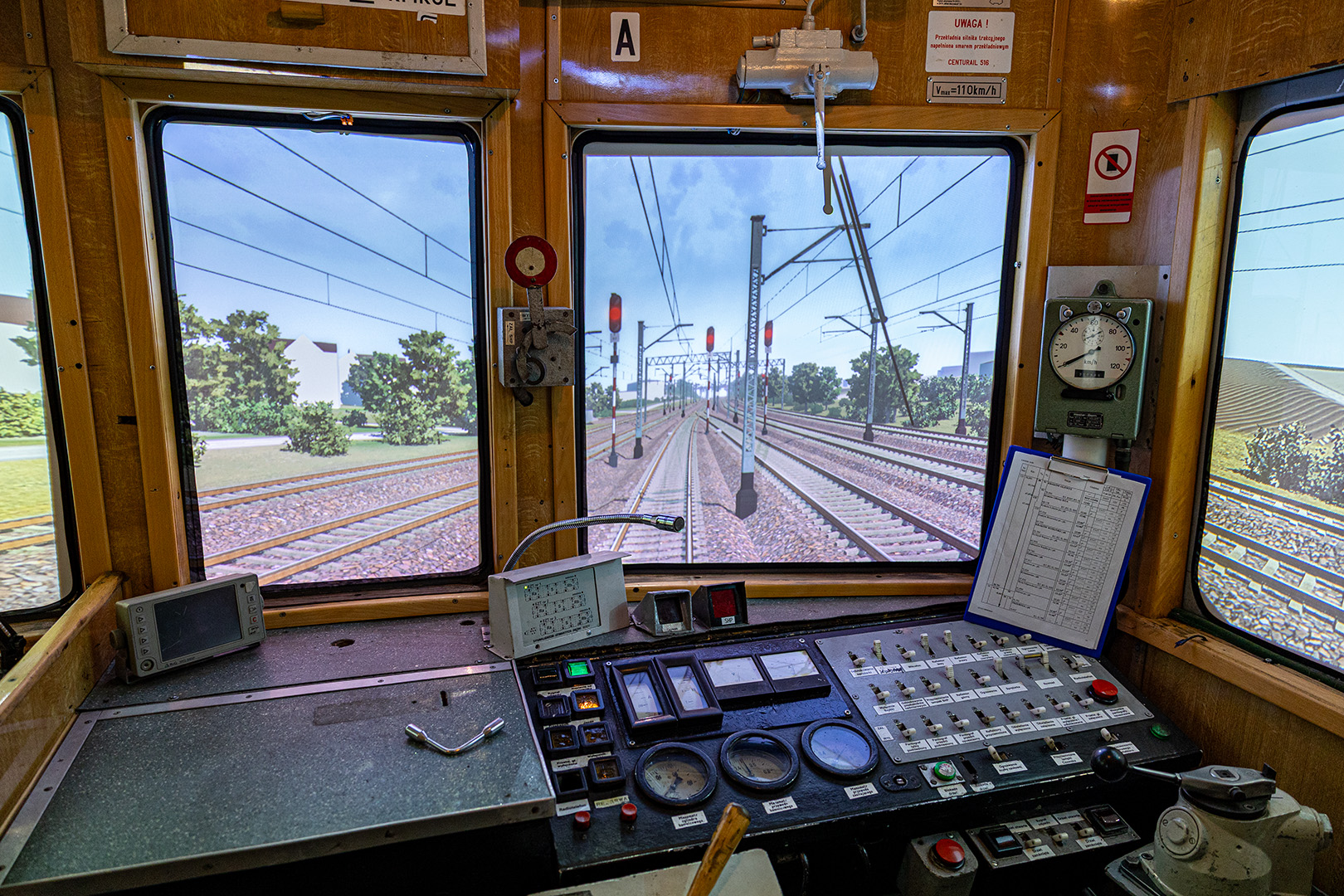 Wnętrze kabiny maszynisty symulatora pociągu elektrycznego. Przed pulpitem sterowniczym z licznymi przełącznikami, wskaźnikami i manometrami widać trzy duże ekrany prezentujące realistyczny widok torów kolejowych z siecią trakcyjną i sygnalizacją świetlną. Widoczne są czerwone światła semaforów oraz słupy trakcyjne wzdłuż torów. Po prawej stronie pulpitu znajduje się tablica z rozkładem jazdy.