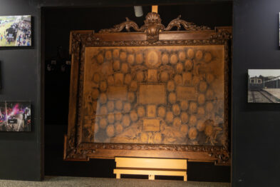 Stare, ozdobne drzewo genealogiczne oprawione w bogato rzeźbioną drewnianą ramę. Wewnątrz widoczne są liczne owalne portrety osób, ułożone w strukturę przypominającą genealogiczne drzewo rodzinne. Całość ma wyblakły, sepiowy odcień, a obraz jest ustawiony na sztaludze w ciemnym pomieszczeniu