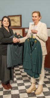 Dwie kobiety stoją w pomieszczeniu o biało-czarnej szachownicy na podłodze i bordowo-niebieskich ścianach, na których wiszą oprawione szkice i zdjęcia. Kobieta po lewej, w czarnym żakiecie i spódnicy, podaje rękę kobiecie po prawej, ubranej w jasny garnitur. Obie trzymają na wieszakach ciemnozielone mundury wyjściowe z ozdobnymi patkami na kołnierzach.