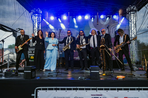 Grupa muzyków i organizatorów stoi na scenie podczas jubileuszowego wydarzenia. W tle widoczne są reflektory sceniczne, instrumenty oraz ekran z grafiką jubileuszu Stacji Muzeum.”
