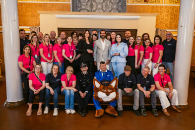 Grupa organizatorów, pracowników i gości jubileuszowego wydarzenia pozuje do wspólnego zdjęcia we wnętrzu zabytkowego budynku. Na pierwszym planie znajduje się maskotka rysia w kolejarskim mundurze.