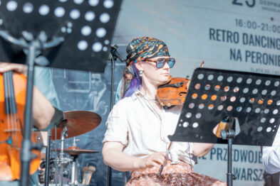 Skrzypaczka w kolorowej chuście i okularach przeciwsłonecznych gra na scenie podczas koncertu zespołu Retro Dancing Band. W tle widoczne są elementy sceny i baner wydarzenia „Niedziela na Głównym”.