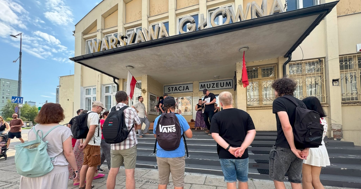 Grupa uczestników spaceru miejskiego stoi przed wejściem do Stacji Muzeum w Warszawie. Przewodnik prowadzi krótkie wprowadzenie na schodach pod charakterystycznym zadaszeniem dawnego Dworca Głównego z widocznym napisem „Warszawa Główna”. Jest słoneczny dzień, a uczestnicy słuchają opowieści o historii warszawskiej kolei i architektury.